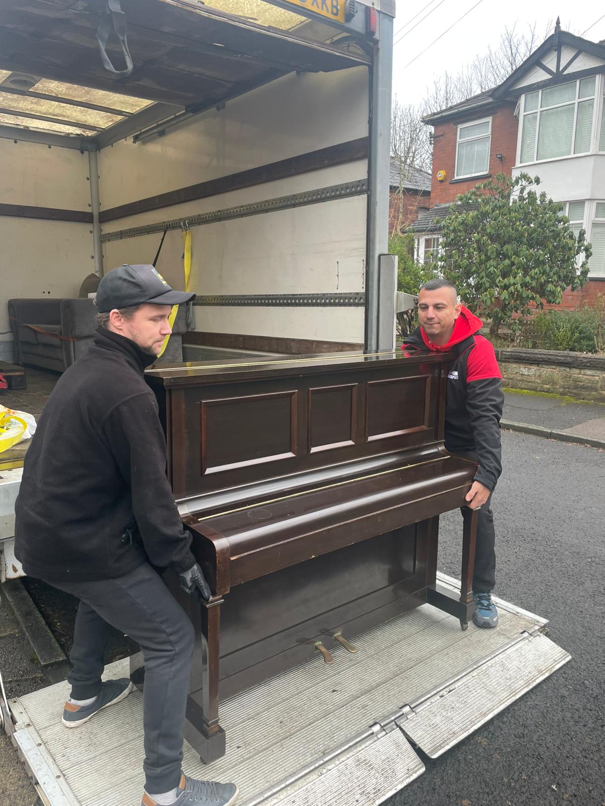 Two Wigan Man & Van movers carefully loading a dark upright piano onto the tail lift of a Luton van
