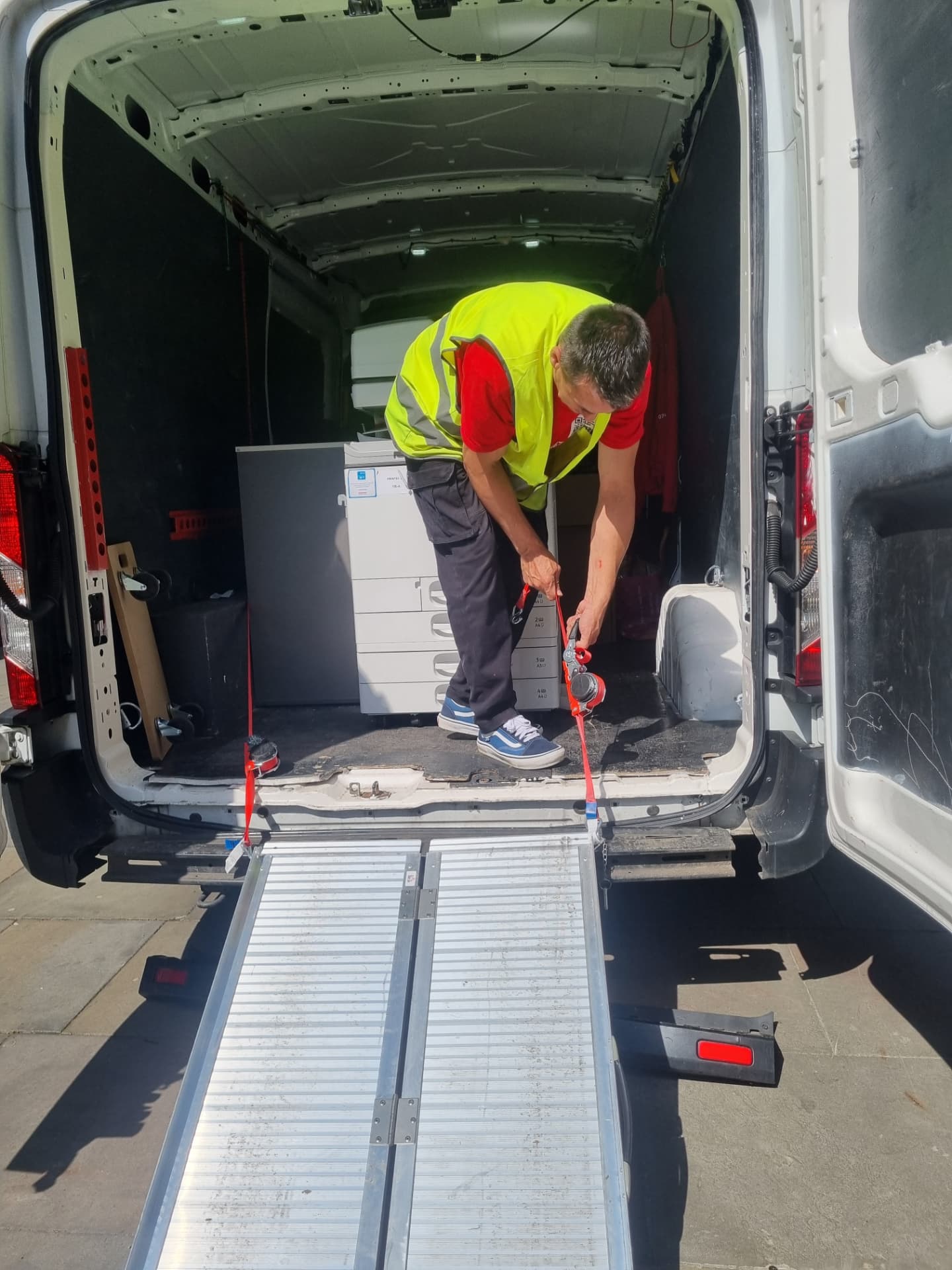 Mover in a hi-vis vest ratchet-strapping an office printer inside the van with the loading ramp down
